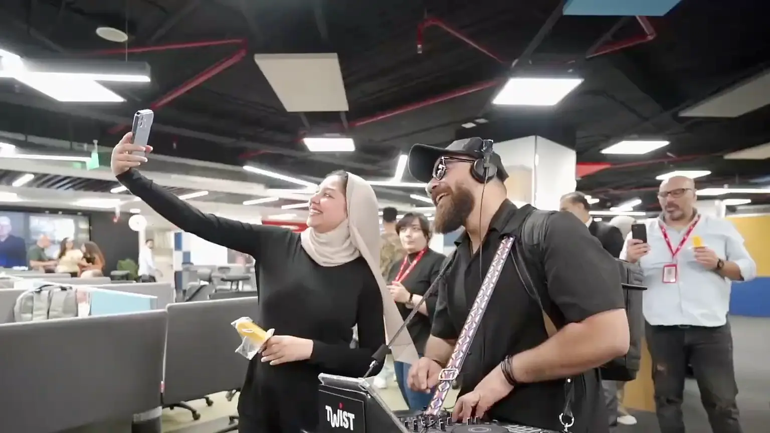 Smiling colleague taking a selfie with a roaming DJ as coworkers film in the open office