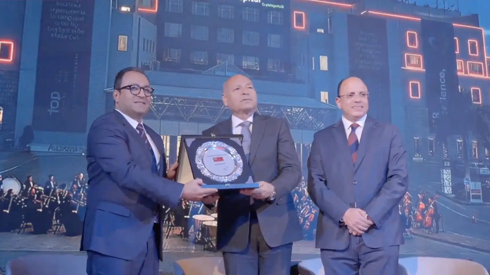 Three senior leaders hold up the award plaque together in front of the stage screen