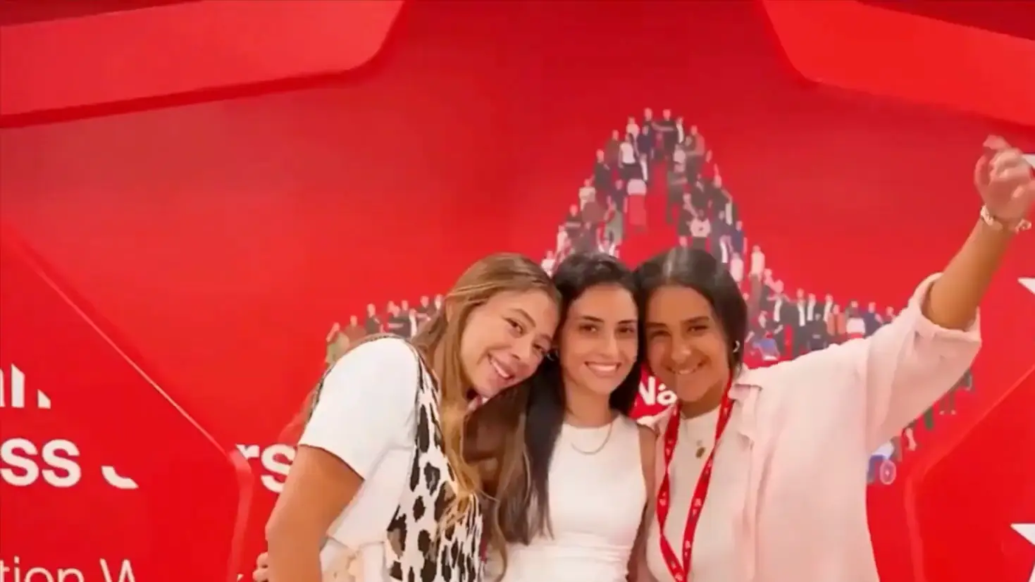 Three smiling young women pose together in front of a bright red employer photo wall