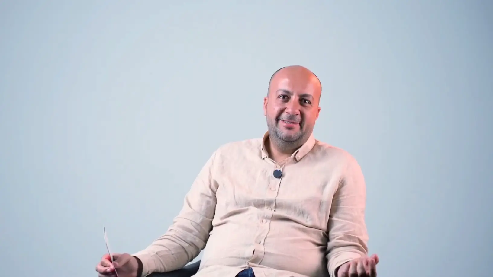 A smiling father in a linen shirt talks openly, seated against a pale studio wall