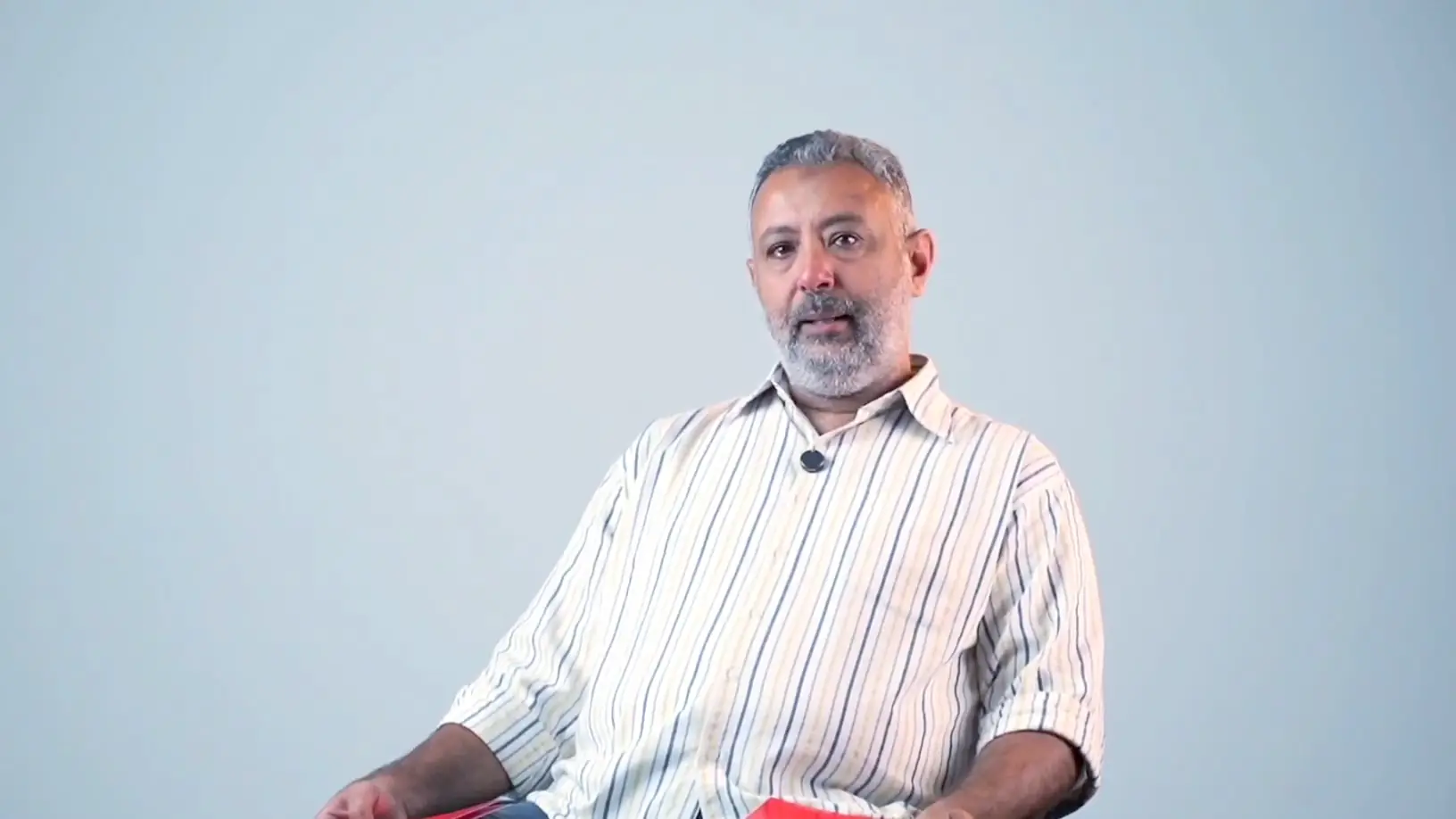 A grey-bearded father in a striped shirt speaks thoughtfully, seated in an interview chair