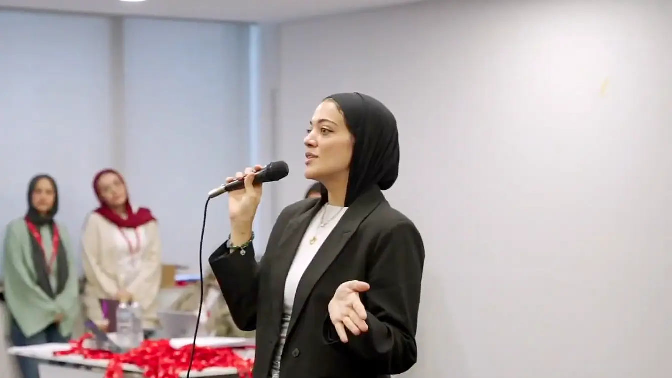 Confident woman in a black blazer speaking into a microphone to a room of interns