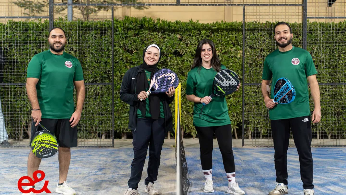 Four colleagues in team shirts holding rackets on a padel court during an evening tournament